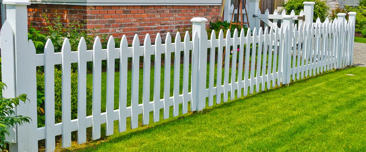A small picket fence on a green patch of grass with a brick home